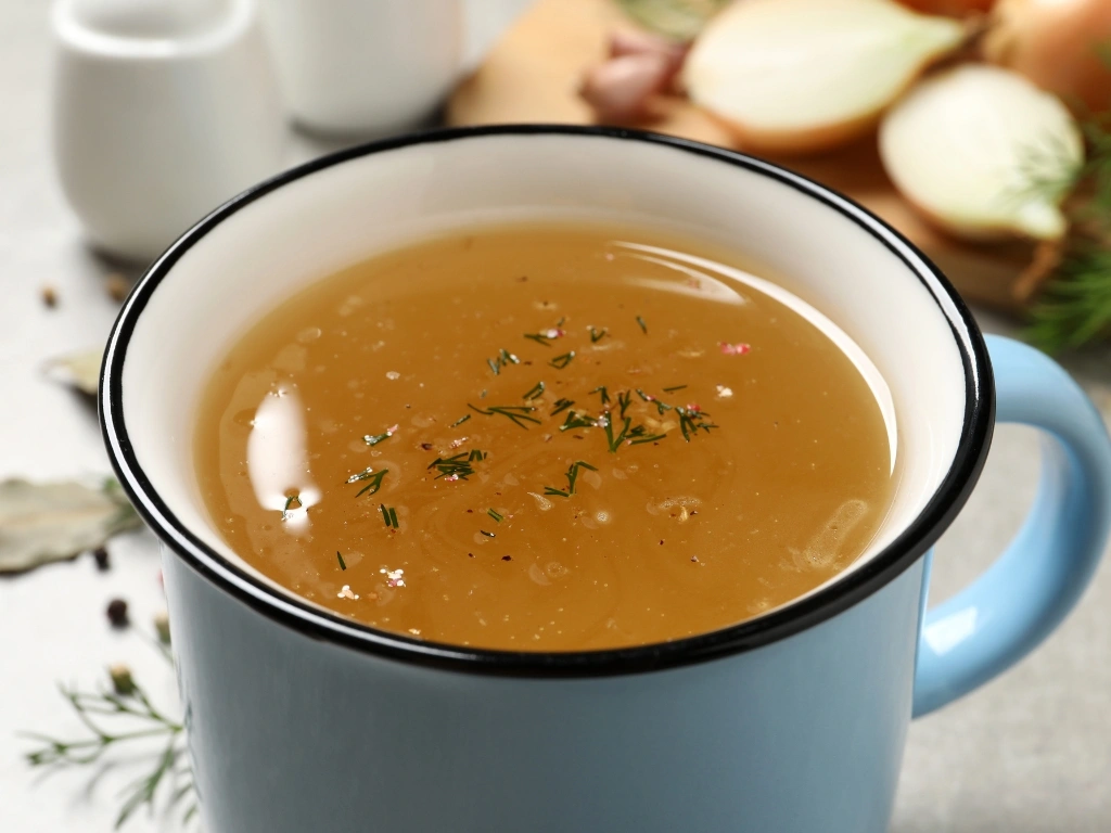 Blue mug filled with savory broth sprinkled with fresh dill, with onions, garlic cloves, sprigs of fresh dill, and white salt and pepper in the background