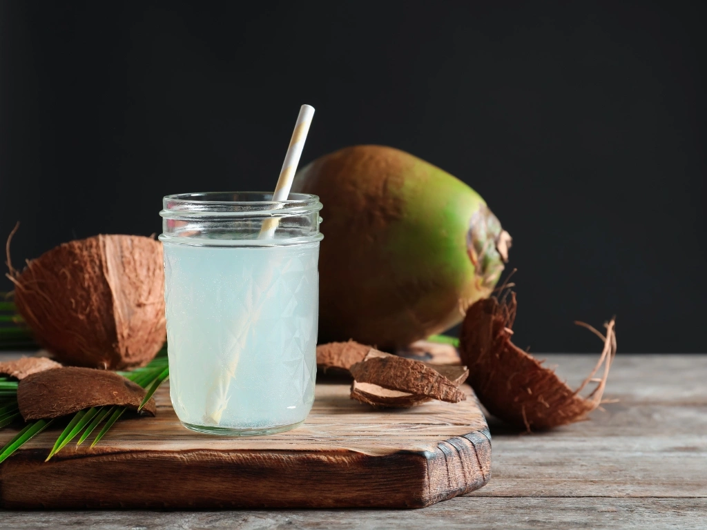 Glass of coconut water served with a straw on a wooden board next to a green coconut and pieces of broken brown coconut shells