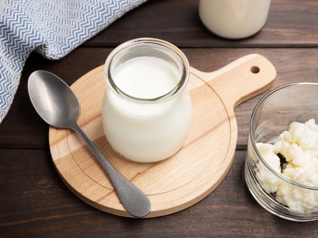Small jar of kefir drink served on a wooden board next to a spoon, a bowl of kefir grains, and a blue tablecloth