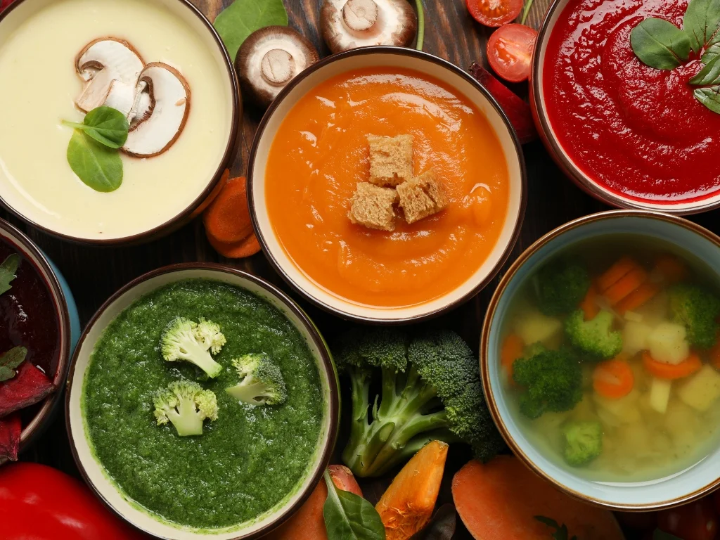 Various bowls of different vegetable soups arranged on a wooden surface next to key ingredients