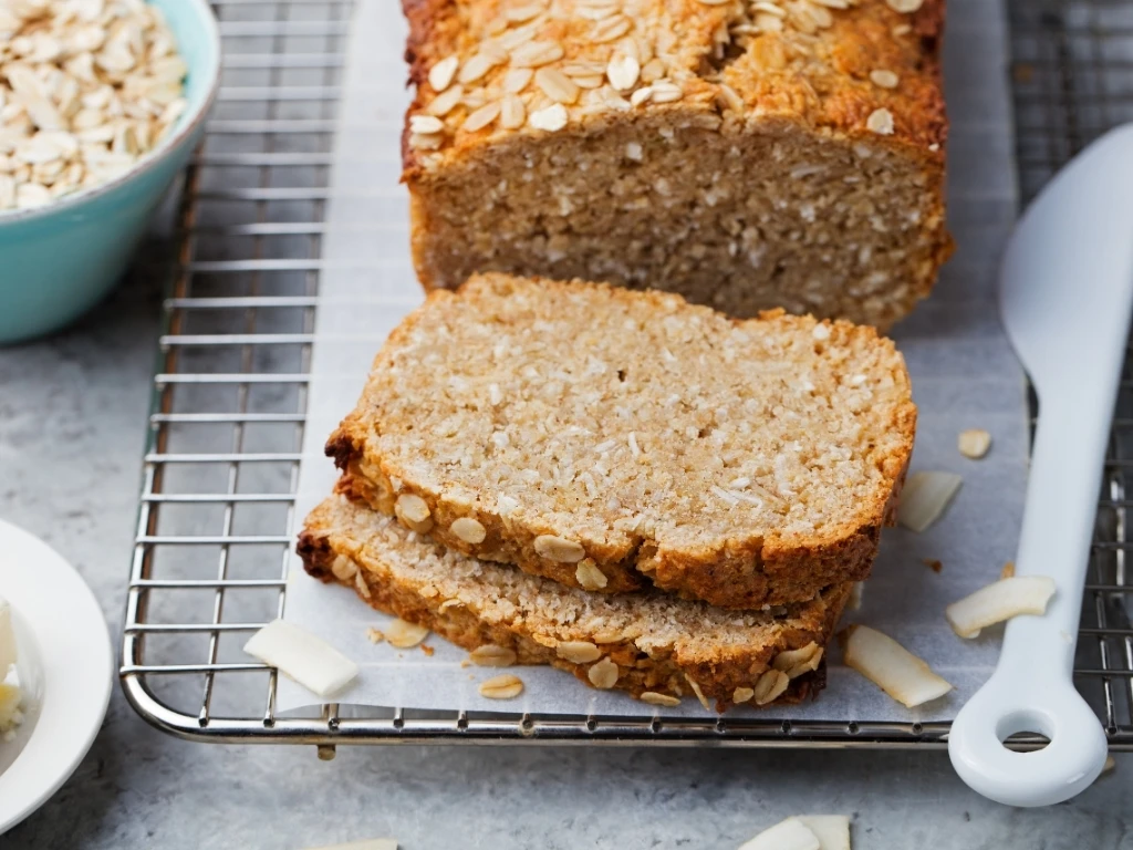 Sliced oat and coconut bread on a cooling rack, next to a white knife, a bowl of uncooked oats, and a small plate of butter