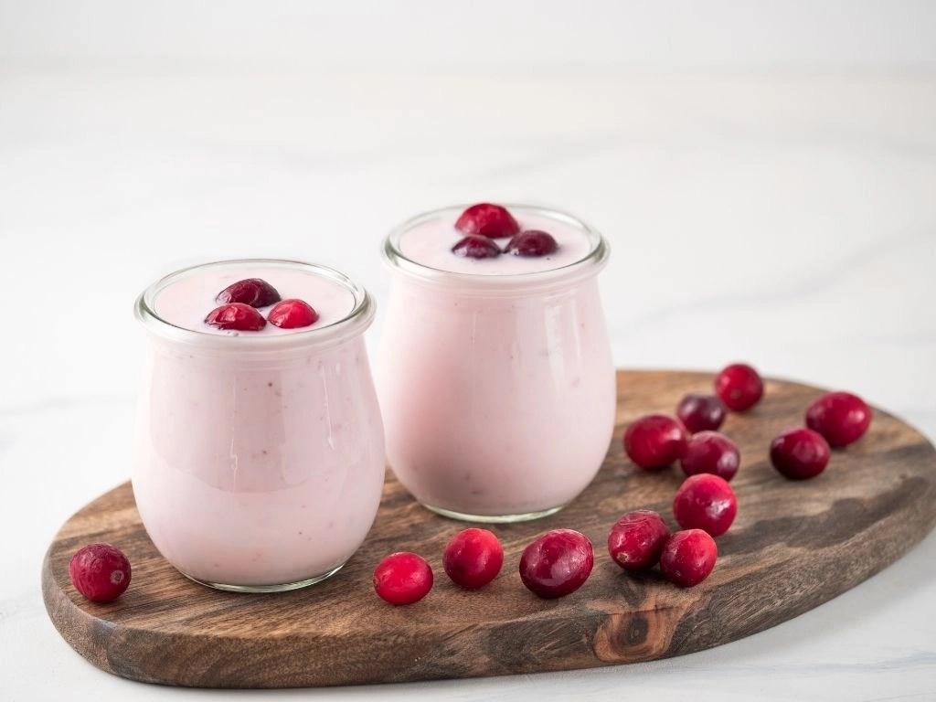 Yogurt with cranberry. Two glass jar with pink yoghurt or milkshake and red cranberries on wooden cutting board over white marble background