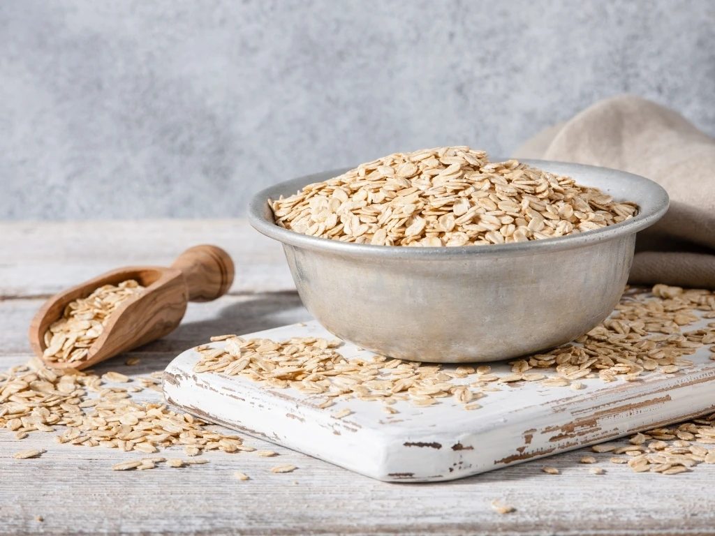 Oats in a metal bowl, with extra oats on a grain scoop and scattered across a wooden board and table