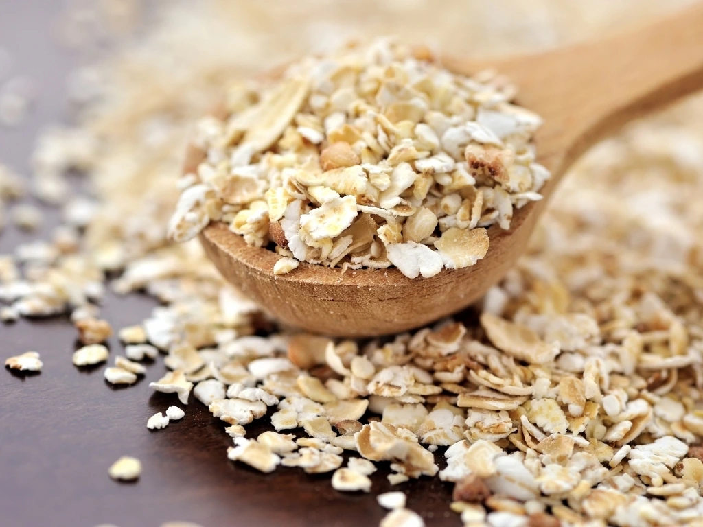 Oats in a metal bowl, with extra oats on a grain scoop and scattered across a wooden board and table