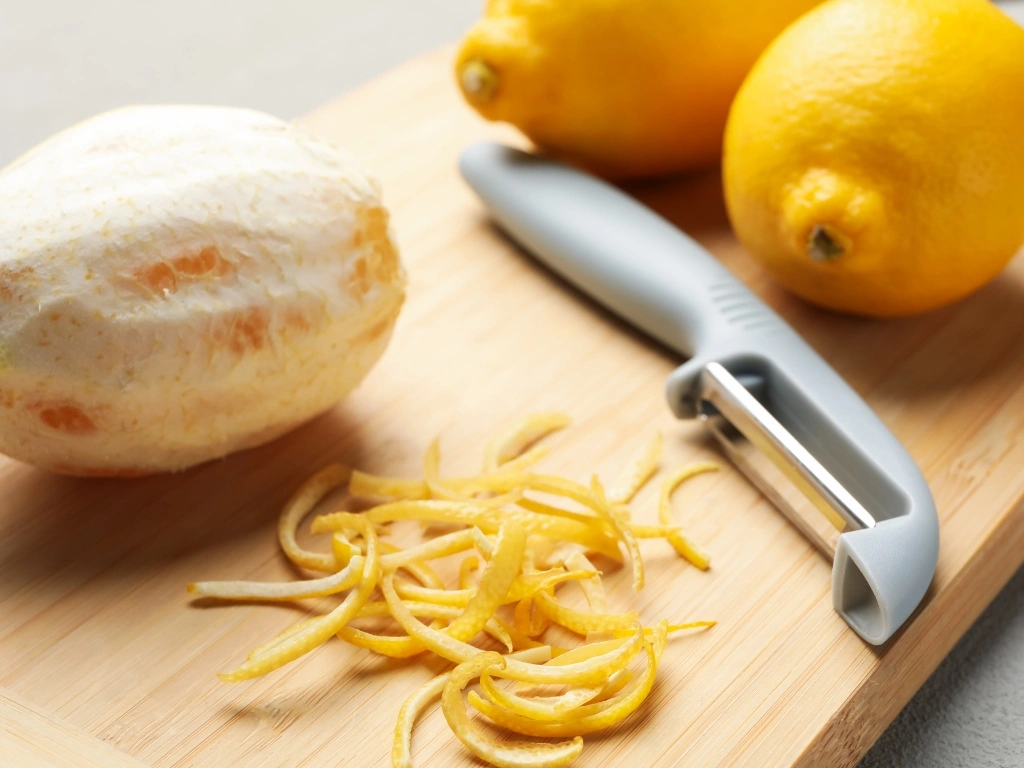 Two whole lemons, a peeled lemon, and strips of zest on a wooden cutting board with a peeler