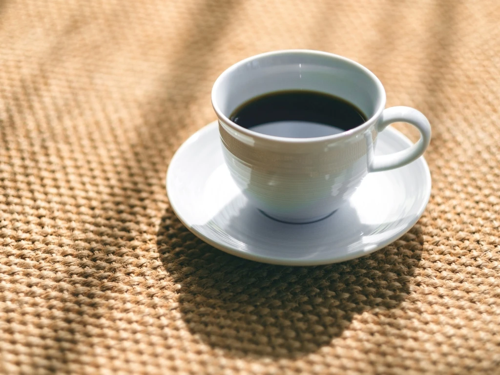 White ceramic cup of black coffee on a matching saucer, resting on a woven brown burlap surface