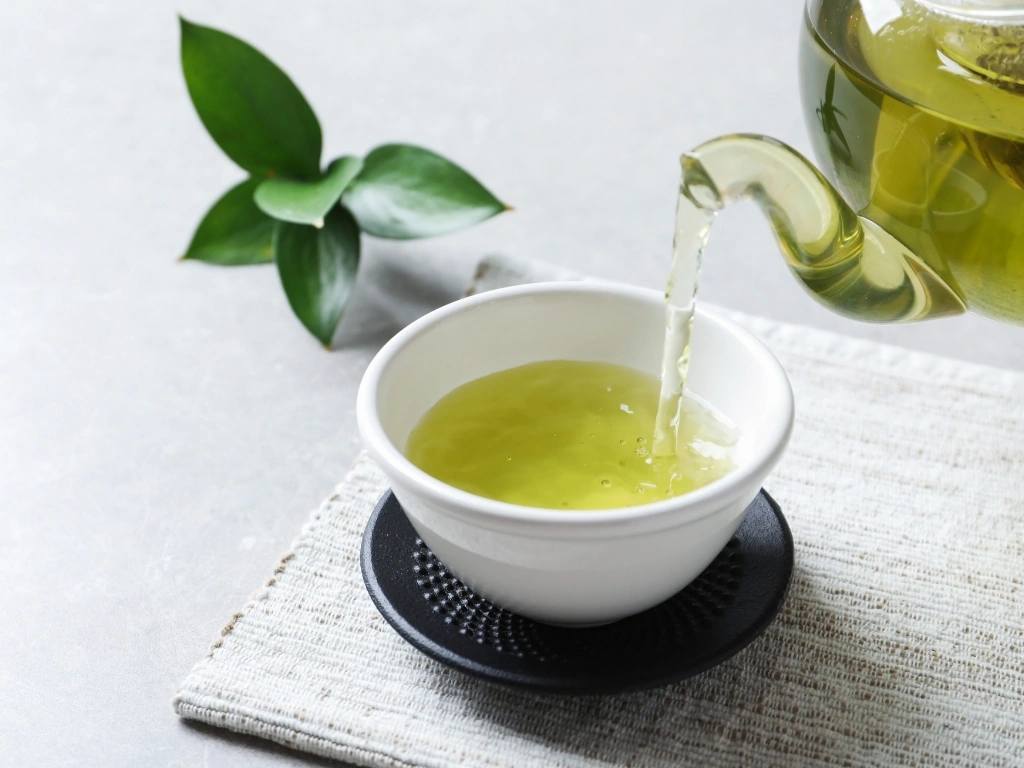Green tea pouring from a glass teapot into a white ceramic cup on a textured black saucer