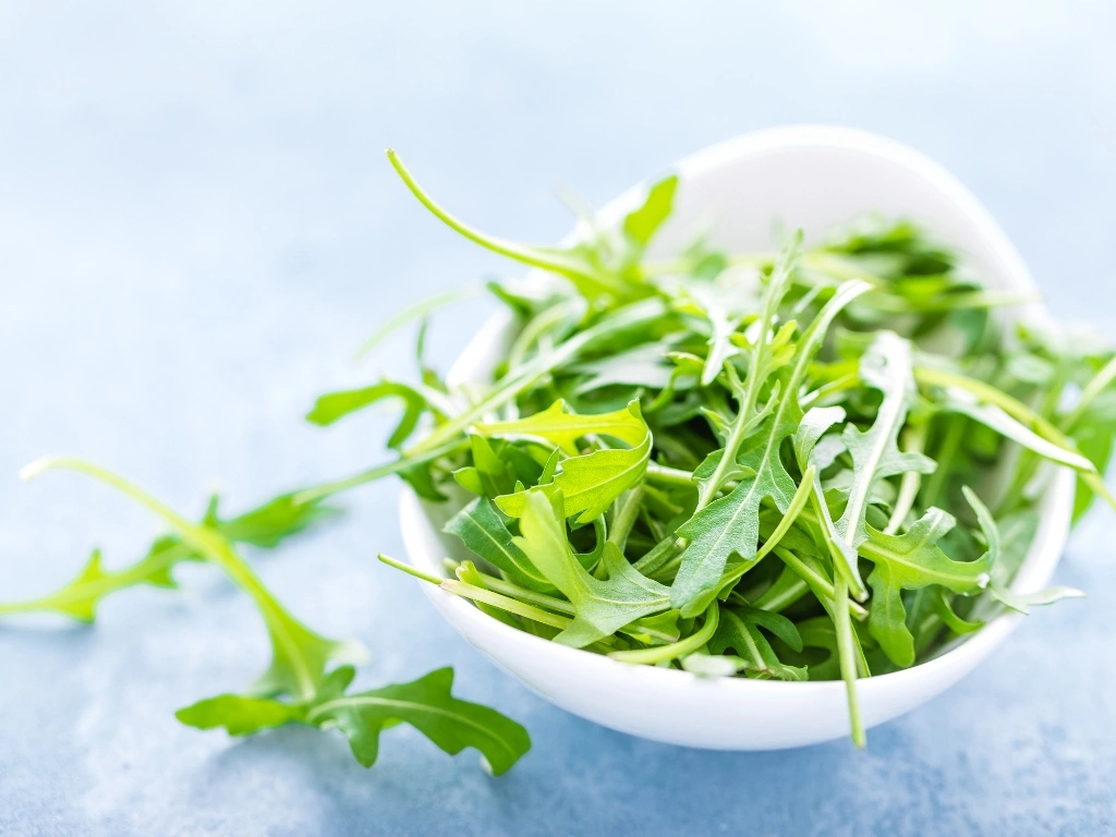 Bowl of vibrant green arugula leaves, set against a light blue textured background