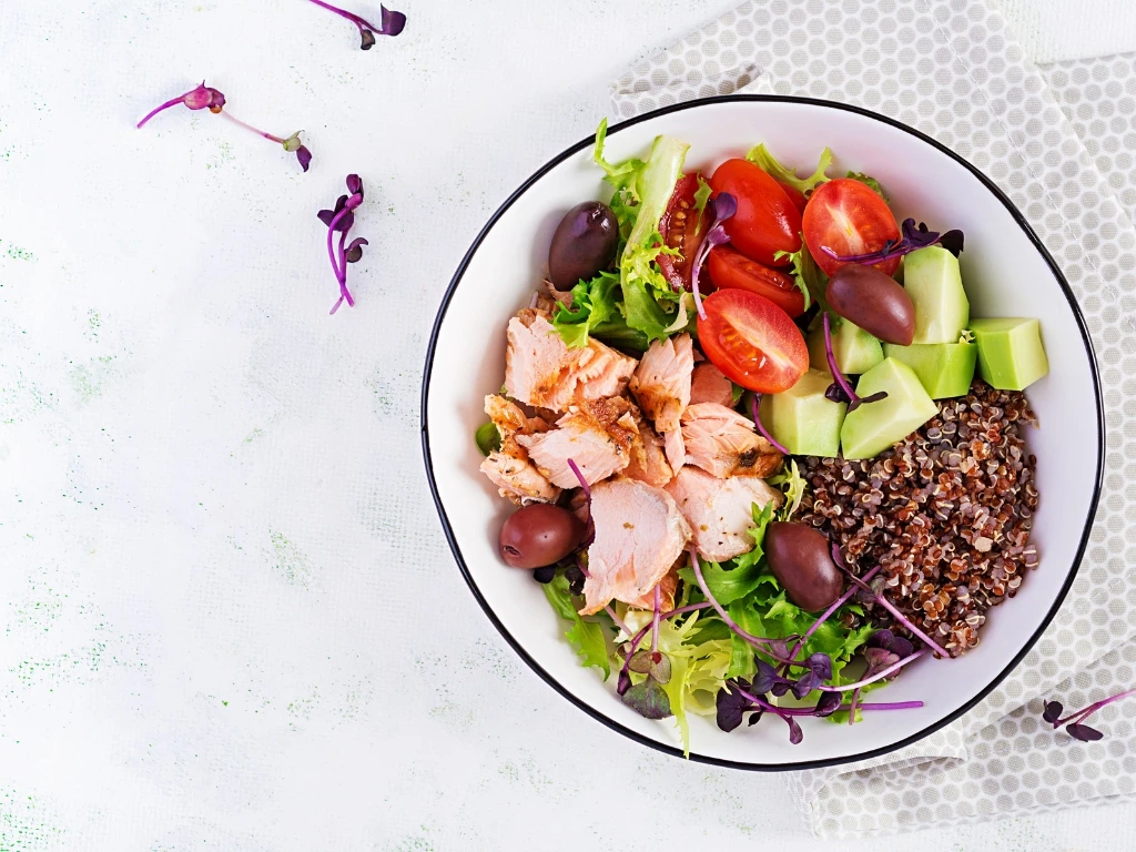 Salmon grain bowl with quinoa, vegetables, olives, and avocado, served on a white plate