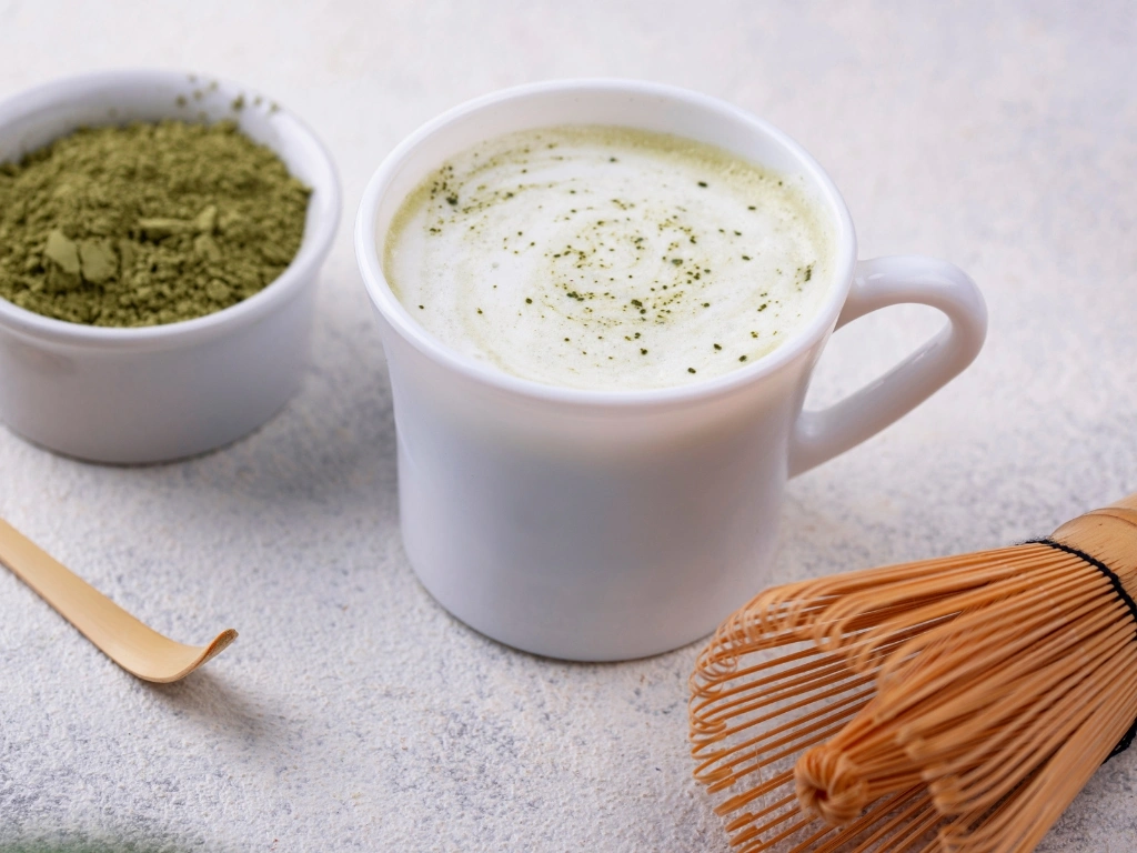 White mug filled with matcha latte, next to a small white bowl filled with matcha powder, a traditional bamboo whisk, and a bamboo spoon