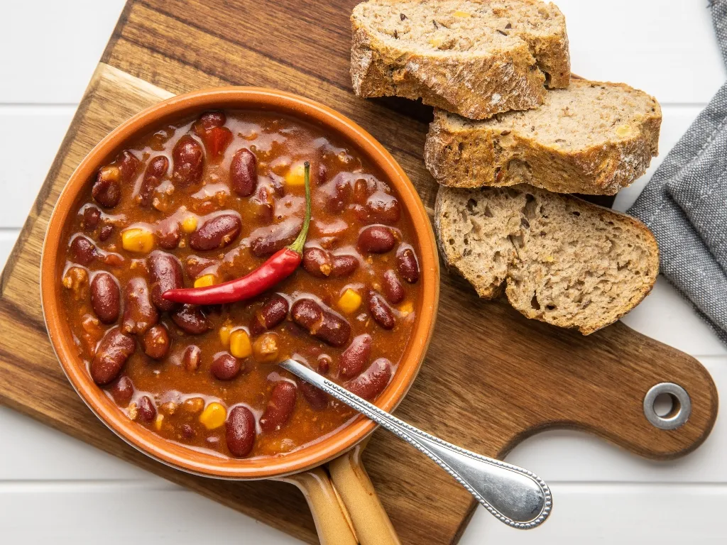 Pot of bean chili served with a spoon on a wooden table, next to slices of whole-grain bread