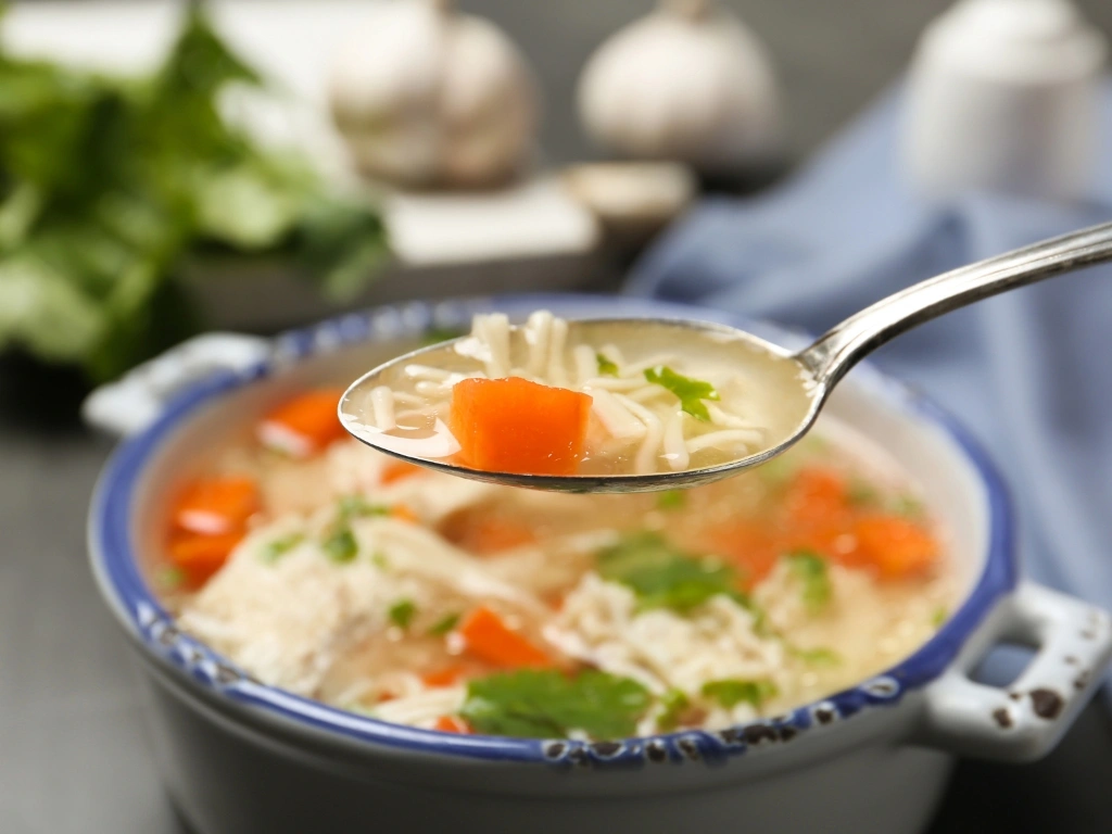 Spoon holding a bite of chicken noodle soup, with the rest of the soup in a blue and white ceramic bowl