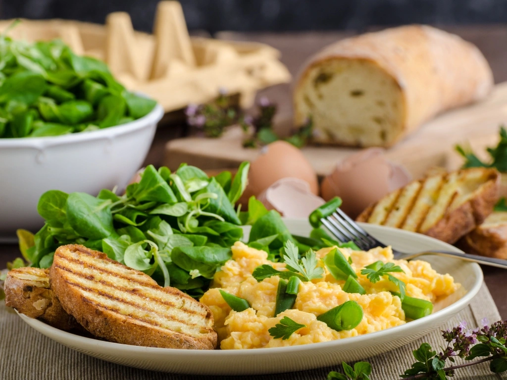 Plate of soft scrambled eggs with mixed greens and toasted bread, with key ingredients served in the background