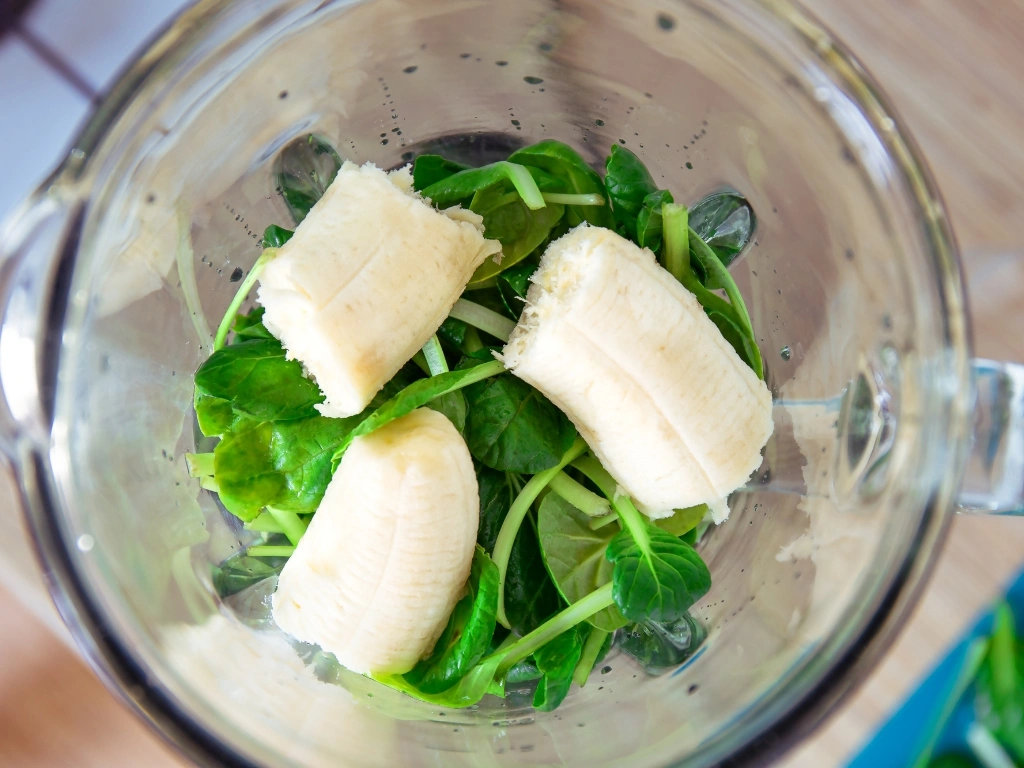 Ingredients for a smoothie in a blender, including banana slices and fresh spinach leaves
