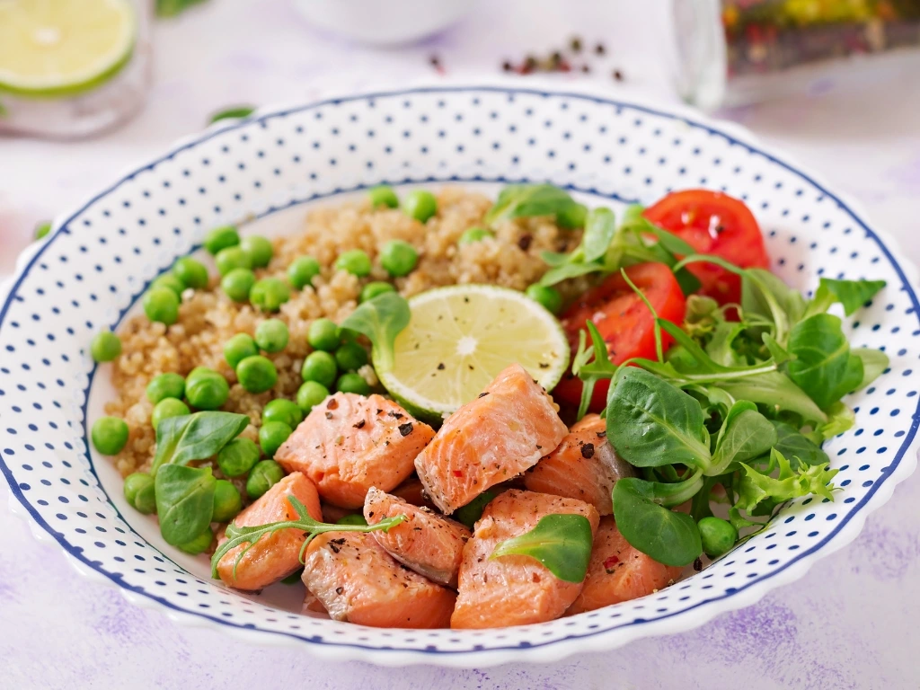 Salmon cubes, quinoa, green peas, tomatoes, leafy greens, and a wedge of lime on a white plate with a blue dotted design