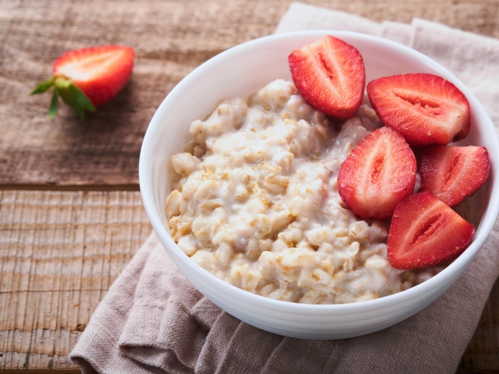 Bowl of oatmeal topped with sliced fresh strawberries, served on a wooden table next to a another sliced piece of strawberry