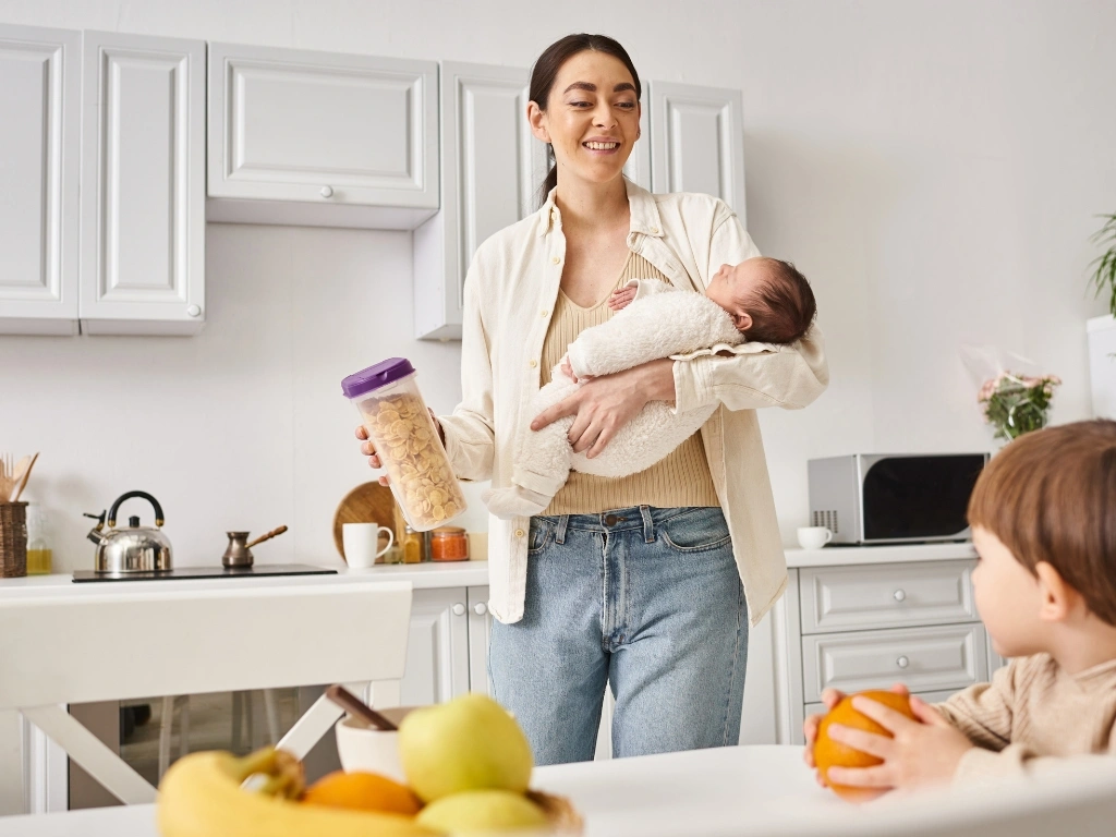 Mother holding a sleeping baby and a cereal container in a kitchen with another child nearby