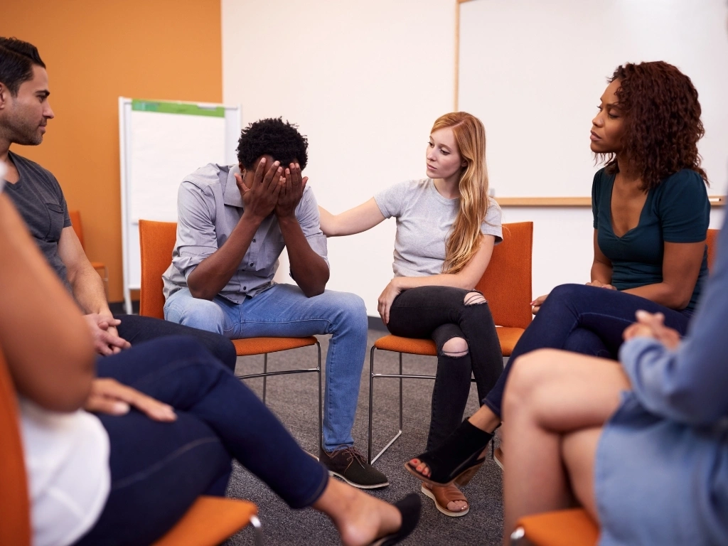 Men and women seated in a circle in a group counseling setting, with one man leaning forward and holding his face