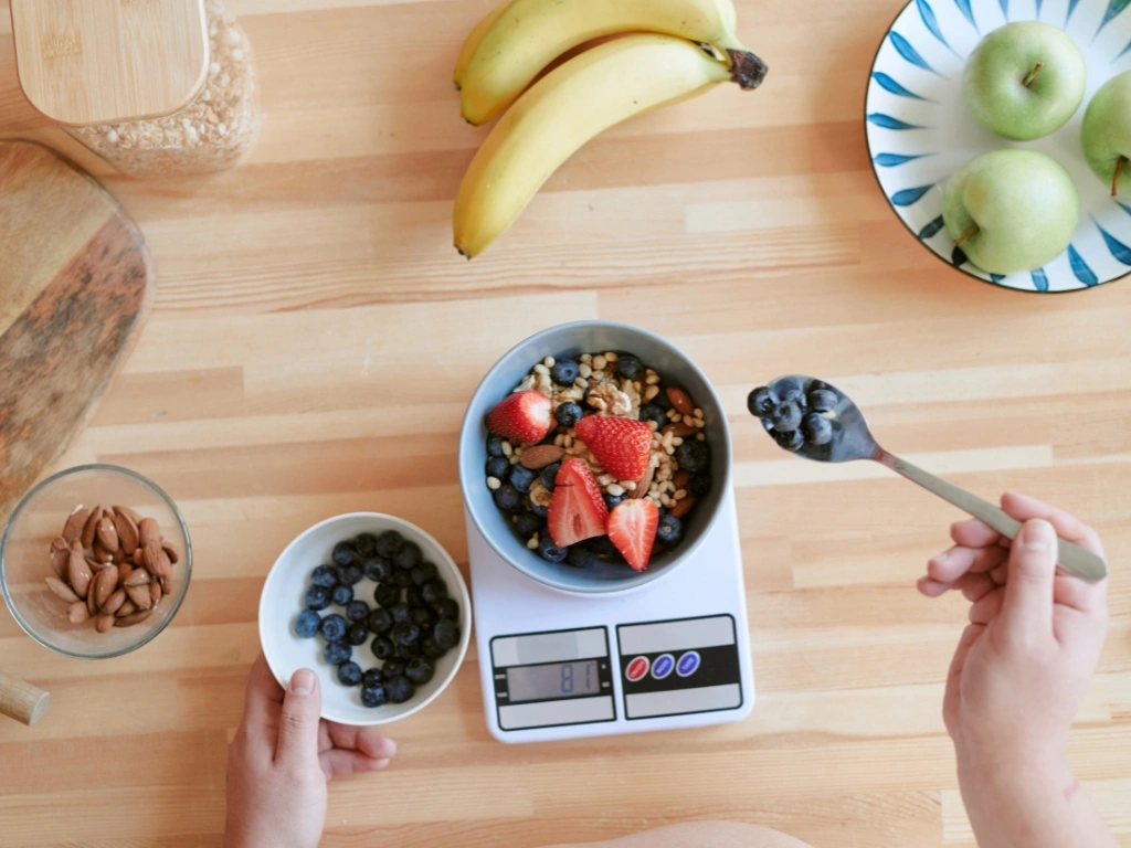 Hands adding blueberries to a breakfast bowl of fruit and nuts on a digital scale