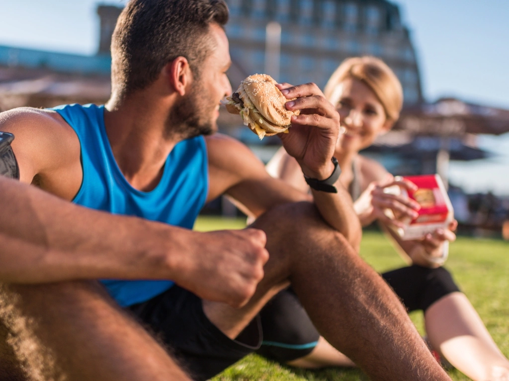 Two individuals in athletic wear sitting on a grassy area while eating burgers