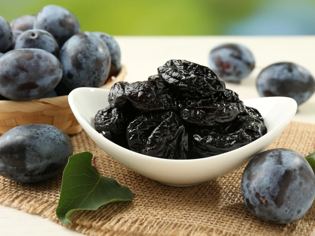 Dried prunes in a white ceramic bowl next to fresh whole plums on a rustic placemat