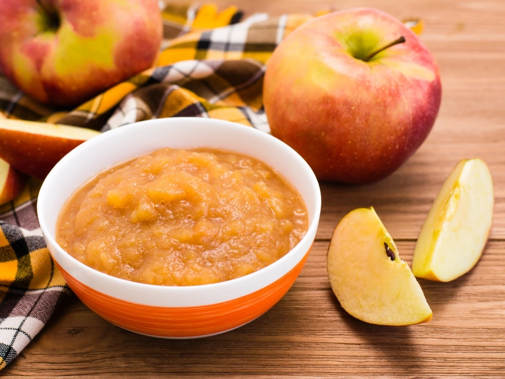 Bowl of fresh applesauce surrounded by whole red apples and apple slices on a wooden table