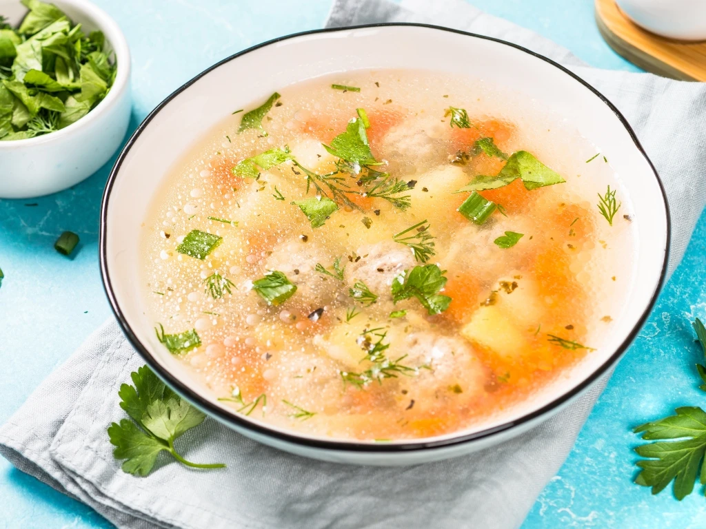 Bowl of vegetable and meatball soup garnished with fresh herbs, served on a blue table next to more herbs