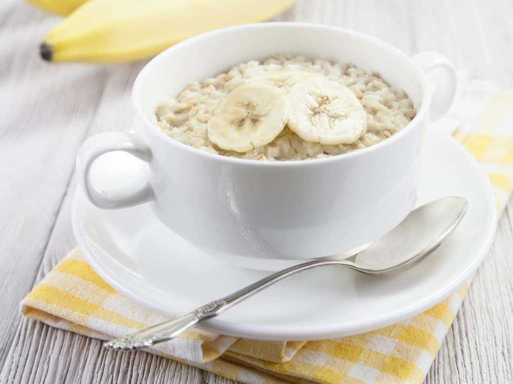 Bowl of porridge topped with banana slices, served in a white cup with a side of whole bananas