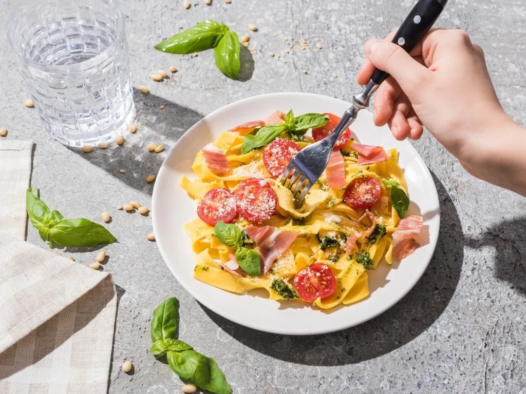 A plate of pasta with tomatoes and prosciutto next to a glass of water