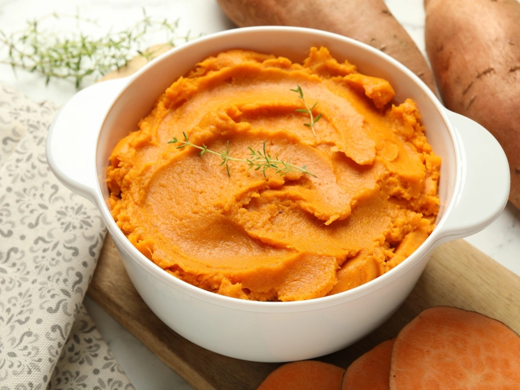 Mashed sweet potato in a white ceramic bowl, served next to raw sweet potatoes and herbs