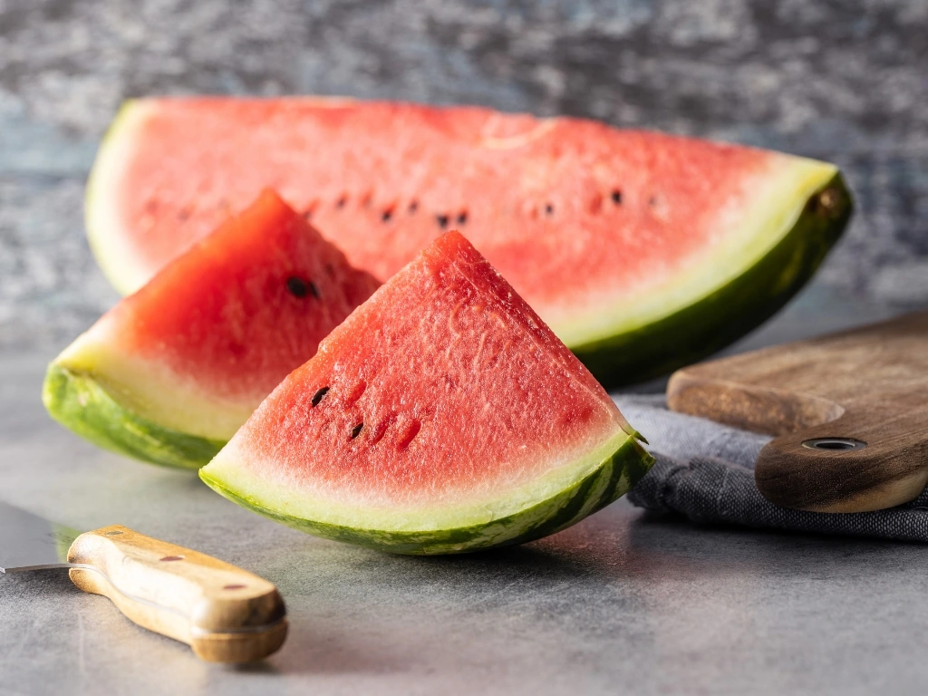 Different sizes of sliced watermelon on a countertop next to a kitchen knife and a wooden cutting board