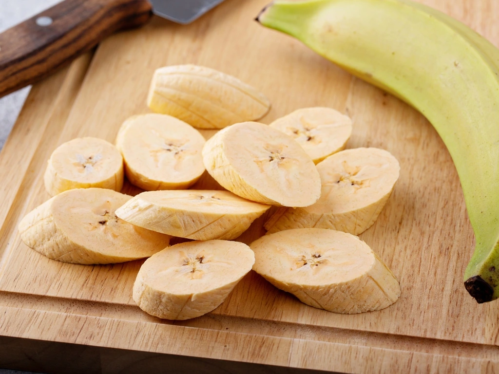 Slices of green banana on a chopping board, next to an unpeeled piece and a kitchen knife
