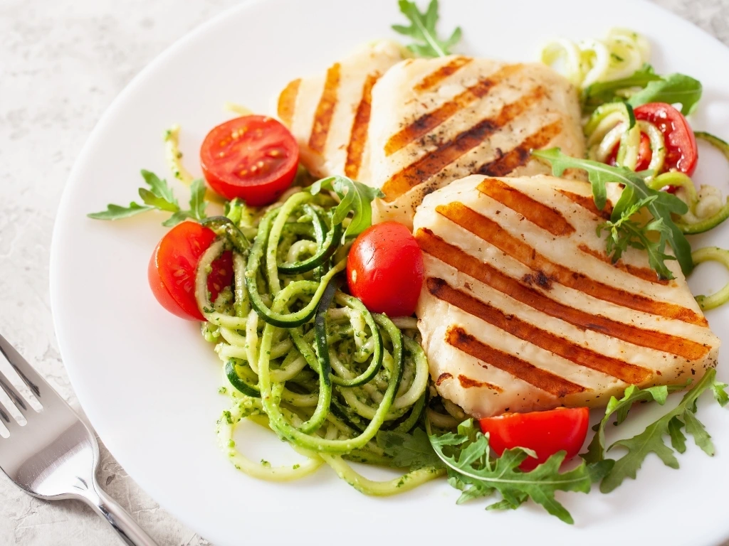 Grilled chicken with zucchini noodles and tomatoes, served on a white plate next to a fork
