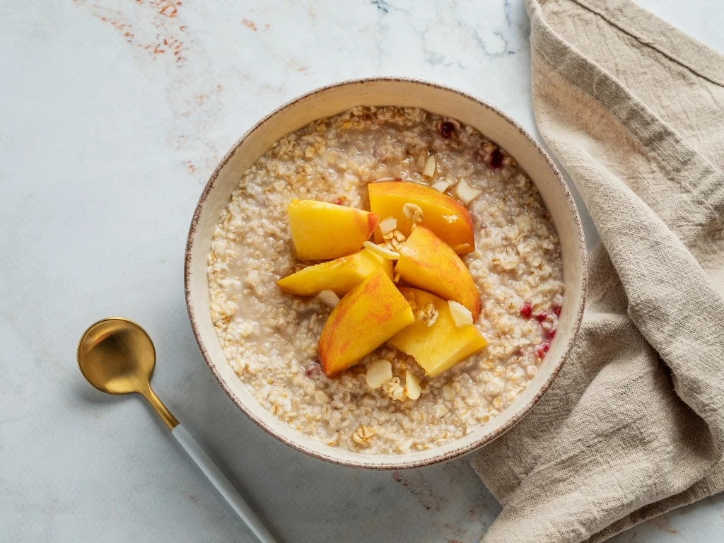 Bowl of oat porridge with peach slices, next to a golden spoon and a tablecloth