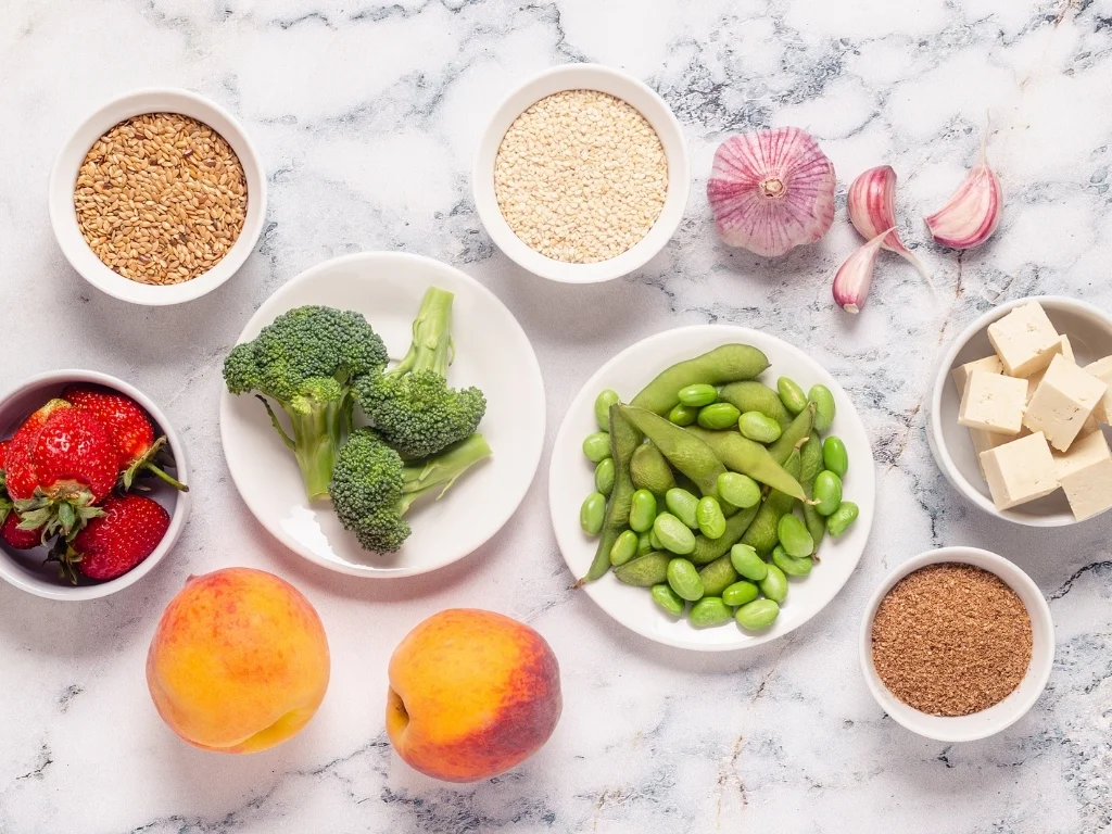 A variety of healthy foods are laid out on a white marble background, including broccoli, peaches, strawberries, tofu, and various seeds and legumes.