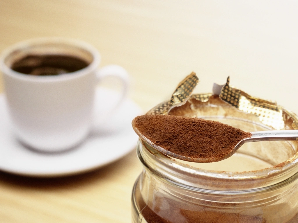 Metal spoon holding instant coffee powder resting on an open glass jar next to a freshly brewed cup of black coffee on a wooden table