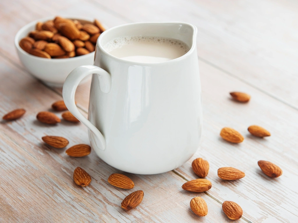 White pitcher filled with almond milk alongside a bowl of whole almonds, set on a wooden table