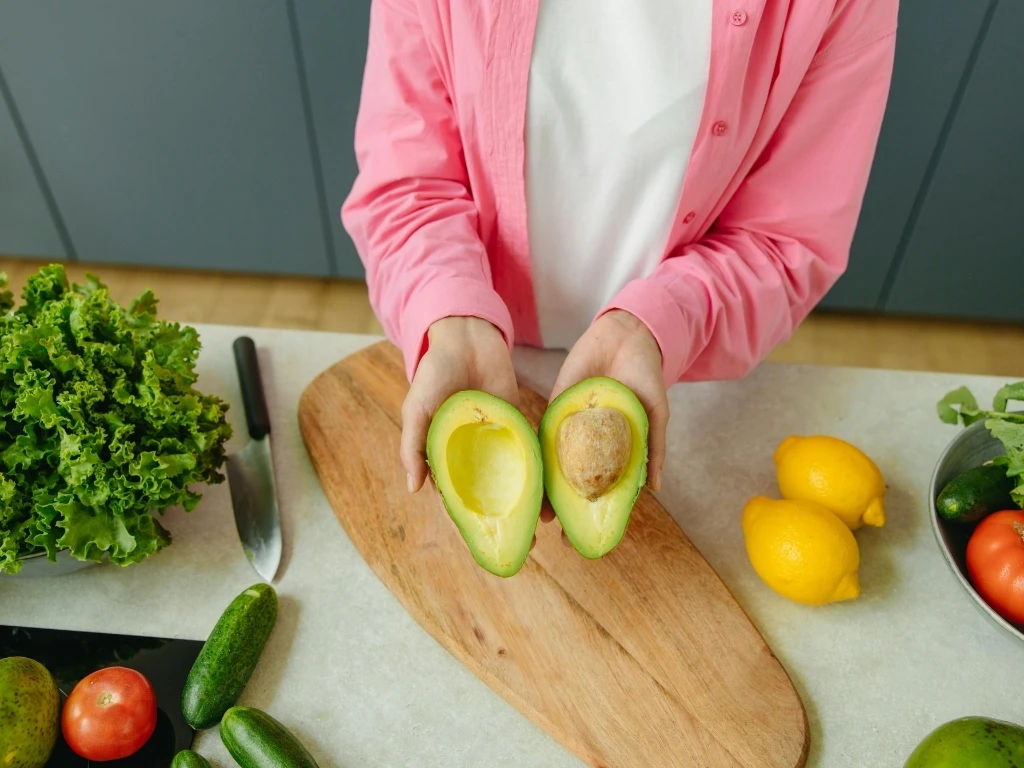 A person holding two halves of a fresh avocado over a wooden cutting board.