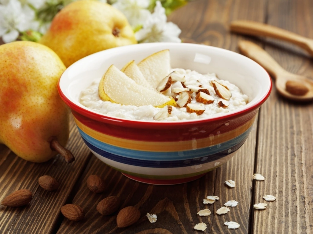 Bowl of creamy oatmeal topped with sliced pears and almonds, next to key ingredients across a wooden table