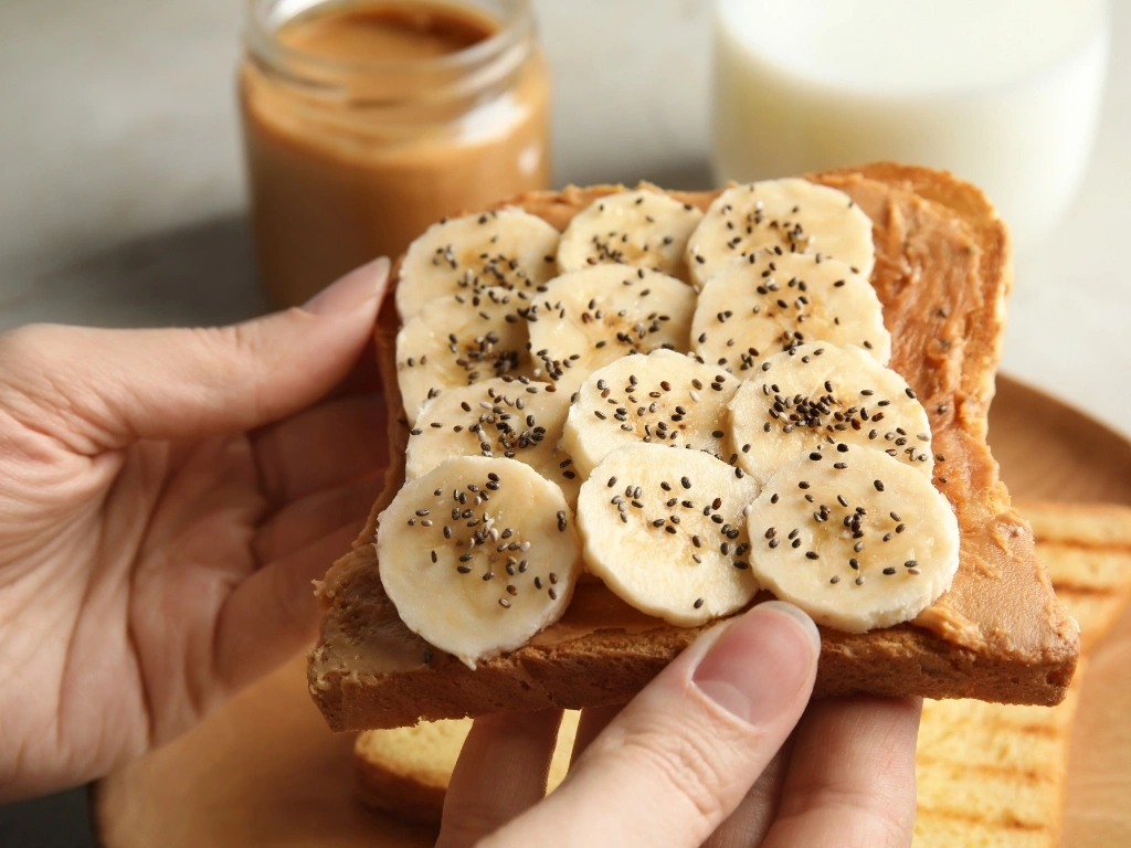 Person holding a slice of whole-grain seeded bread topped with peanut butter, banana slices, and chia seeds