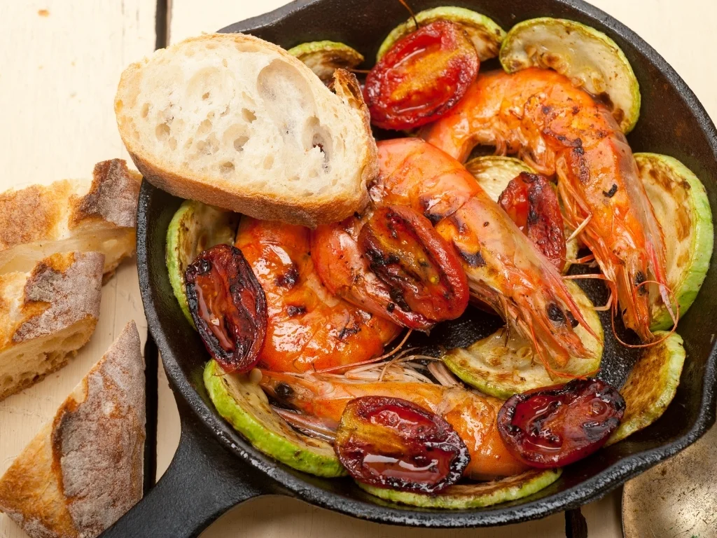 Cast-iron skillet of grilled whole shrimp, roasted tomatoes, and zucchini rounds served with crusty bread slices