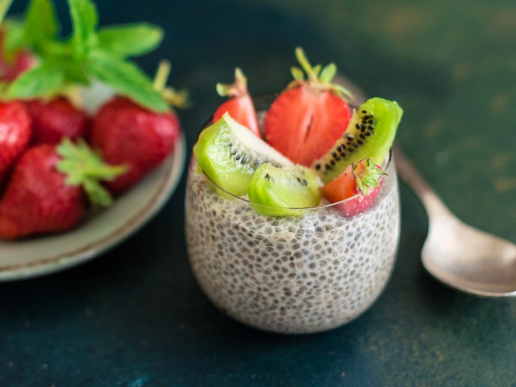 Glass of chia pudding topped with sliced kiwi and strawberries, next to a spoon and a plate of fresh, unsliced strawberries