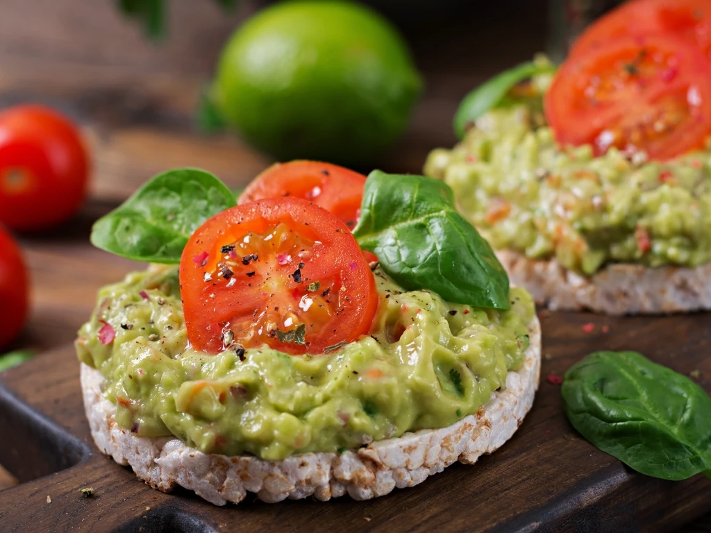 Rice cakes topped with guacamole, sliced tomatoes, and basil on a wooden board, next to key ingredients