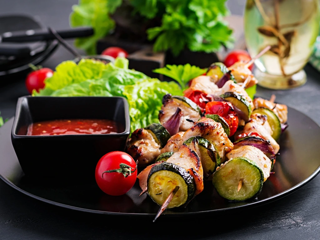 Grilled chicken and vegetable skewers served on a black plate with a red dipping sauce and a side salad