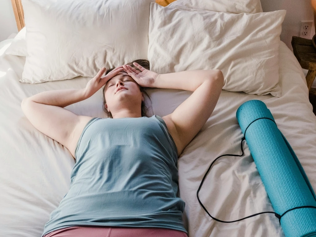 Woman tired from a workout, lying in bed next to a rolled-up yoga mat
