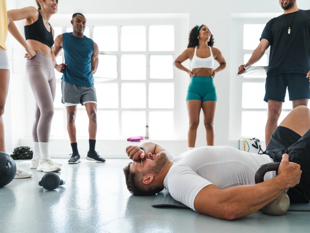 Man lying down on the gym floor in pain while holding a kettlebell, while other gym members stand nearby