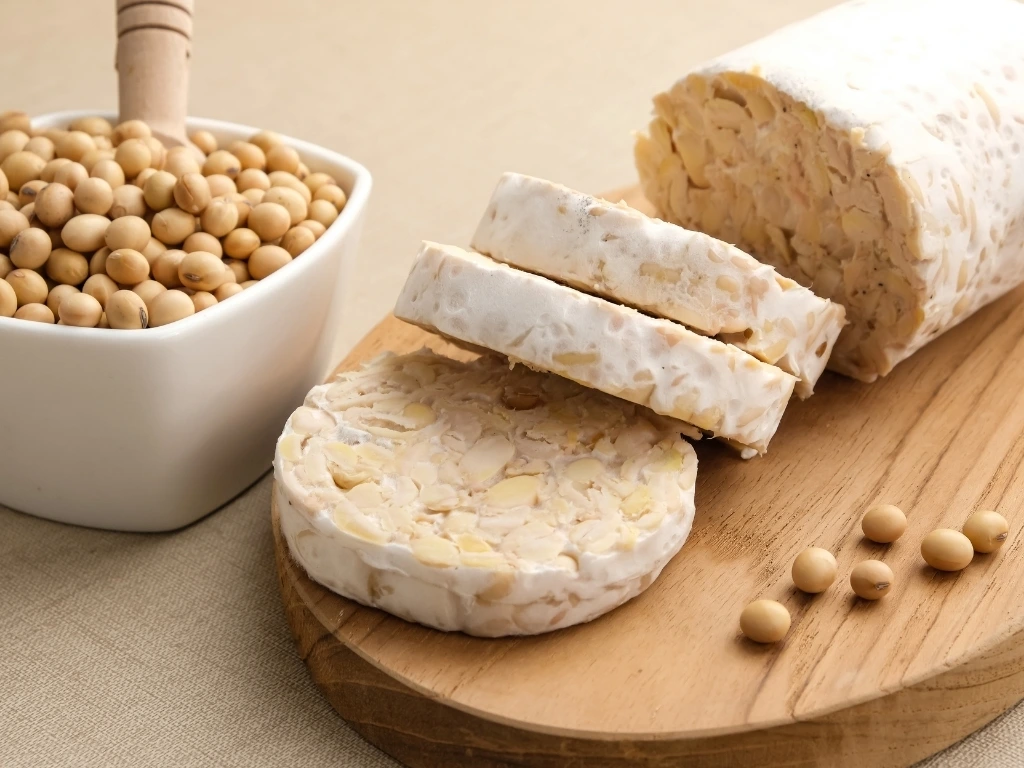 Tempeh slices on a wooden board, next to a container of soybeans