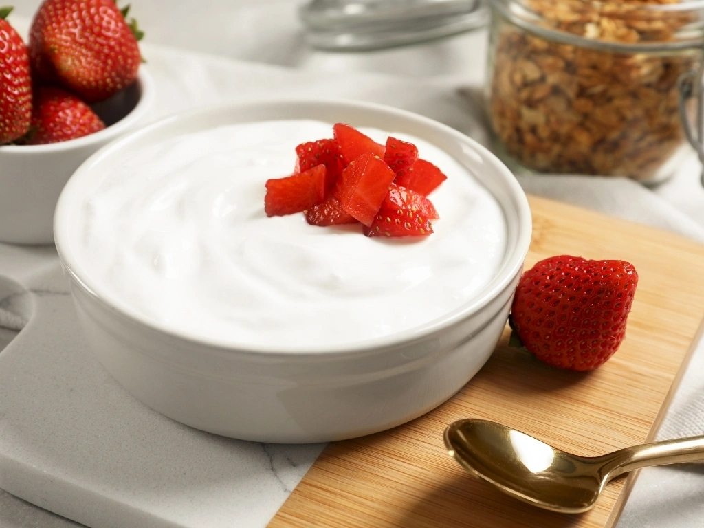 Bowl of yogurt with fresh strawberries, served on a board next to unsliced pieces of the fruit, a spoon, and a jar of granola