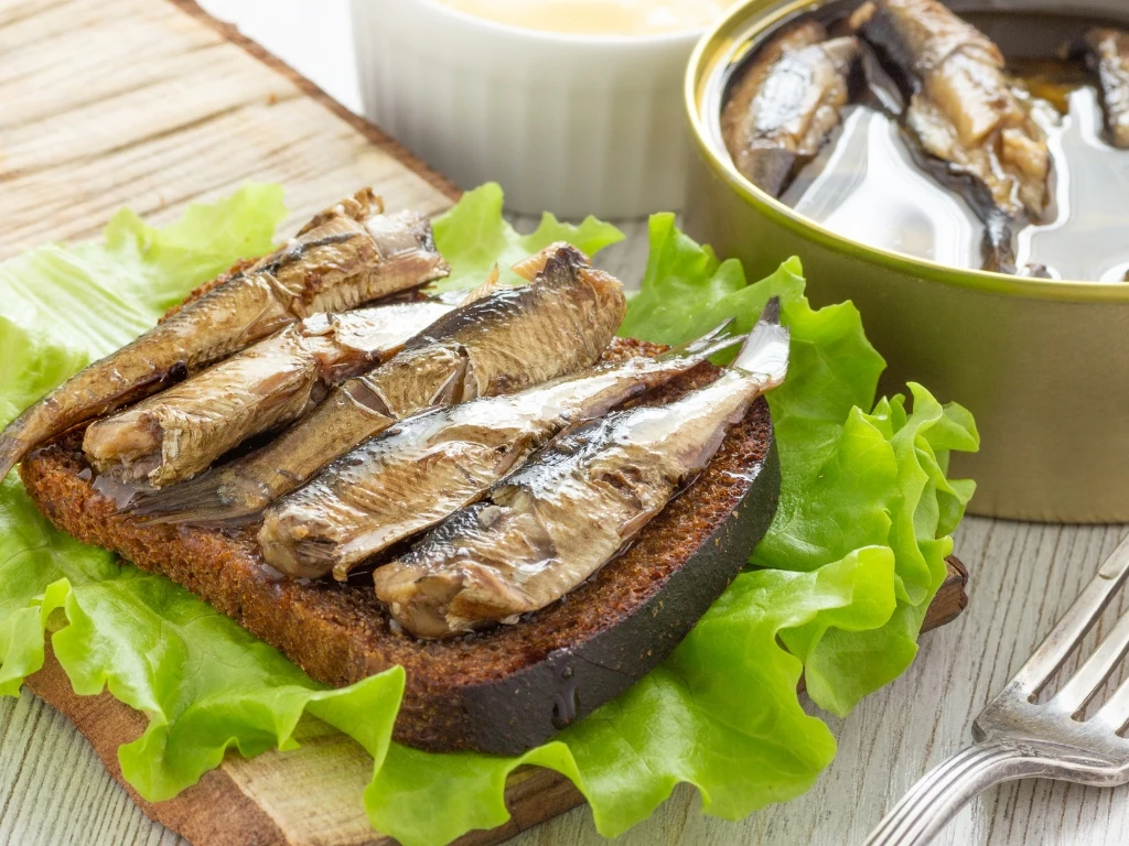 Canned fish on a slice of dark bread resting on green lettuce leaves, next to an open can of sprats and a fork