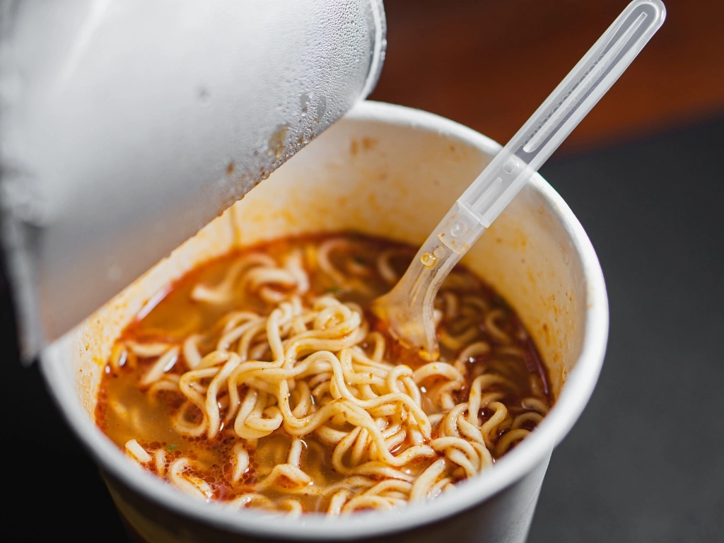 Steaming cup of spicy instant noodles with curly ramen in a red broth and a plastic fork